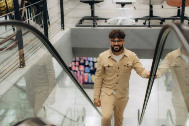 Yürüyen merdivende dururken gülümseyen gözlüklü genç bir adam sıradan bir alışveriş gezisinde eğleniyor.