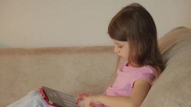 Küçük bir çocuk kanepeye oturur ve tabletinde oyun ya da piyano simülatörü uygulaması oynar. Oynarken gülümsüyor..