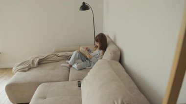 Genç bir kız bej bir kanepede rahatça oturur, bir kitaba dalmış olarak. Samimi ortam ve hafif ışıklandırma okumak için mükemmel bir ortam yaratıyor..