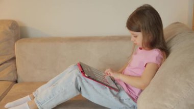 Genç bir kız kanepede oturur ve bir tablette piyano uygulaması çalar. Pembe bir gömlek ve açık mavi kot pantolon giyiyor..