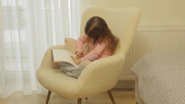 Plush sandalyesinde rahat bir şekilde oturan küçük kız, Pencere kenarındaki defterine çizim yapıyor. Yaratıcılık ve Çocukluk Gelişimi.