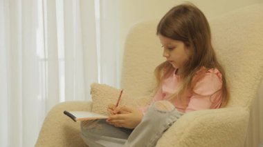 Genç bir kız rahat bir sandalyeye oturur ve defterine mutlu bir şekilde çizim yapar. Çizimini bitirdi, sayfayı yırttı ve gülümsedi..