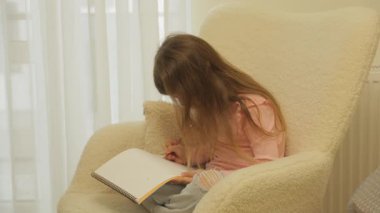 Uzun kahverengi saçlı genç bir kız bej bir koltukta rahat bir şekilde oturuyor, kırmızı renkli bir kalemle boş bir defter çiziyor..