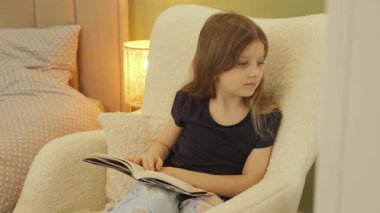Genç bir kız yatak odasındaki rahat bir sandalyeye oturmuş, büyüleyici bir kitaba dalmış. Ilık aydınlatma sıcak bir atmosfer yaratır..