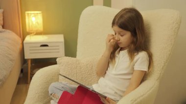 Genç bir kız rahat bir sandalyede oturuyor, tablet kullanıyor. Düşünceli ve ekrandaki içerikle meşgul görünüyor..