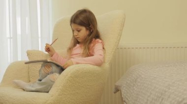 Genç bir kız pofuduk bir sandalyeye rahatça oturur, kırmızı bir kalemle çizim ya da yazıya dalmıştır..