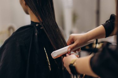 Bir kuaförde, bir müşterinin ıslak, koyu renk saçlarında düz ütü kullanarak profesyonel bir saç tedavisi uygulayan stilistin elleri.