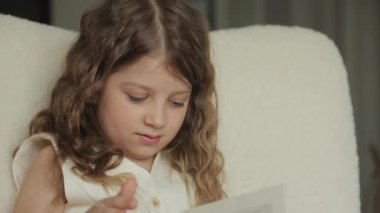 Kıvırcık kahverengi saçlı genç bir kız krem renkli bir sandalyeye oturur ve kucağındaki bir şeye bakar. Gülümsüyor..