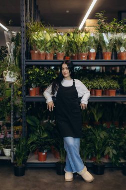 Önlük takan genç bir kadın, bitki yetiştirme odasında dikiliyor ve gülümsüyor çeşitli saksı bitkileriyle çevrili, çalışmalarını sergiliyor.