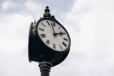 Roma rakamlarıyla süslenmiş sokak saati, siyah eller ve beyaz yüz parlak bulutlu gökyüzüne karşı, zamanı sembolize ediyor.