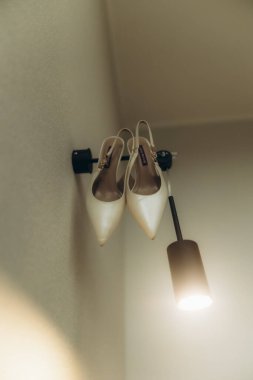 Duvar lambasına asılmış zarif beyaz gelin topukluları düğün törenine hazır. Büyük güne hazırlanıyorum.