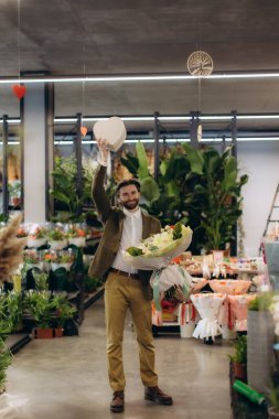 Gülümseyen adam aşkı ve hediyeyi kutluyor, elinde taze bir çiçek buketi ve perakende dükkanında kalp şeklinde bir kutu tutuyor.