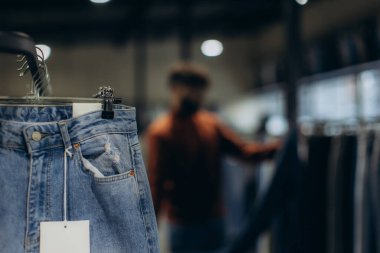 Askıda etiketli mavi kot pantolon, yeni moda kıyafetleri sergileniyor. Arka planda giysileri karıştıran bulanık bir kişi