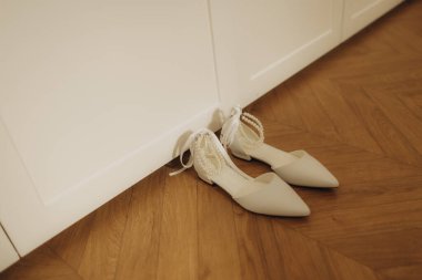 Bir gelin için zarif beyaz düz ayakkabılar, zarif inci bileklikler ve saten kurdeleler, parlak ahşap bir zeminde dinleniyor.