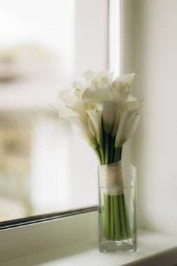 Beyaz zambak buketi parlak bir pencere eşiğindeki cam vazoda duruyor, saflığı ve kutlamayı temsil ediyor.