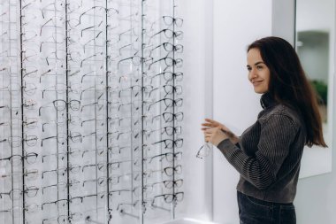 Kadın bir optik mağazada duruyor, vitrindeki çeşitli gözlüklerden bakıyor, görüş düzeltmesi için yeni gözlük düşünüyor.