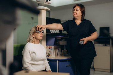 Optisyen, modern oftalmoloji kliniğinde göz muayenesi sırasında deneysel çerçevenin ayarlanması ve kıdemli hastanın görüşünü test ediyor.