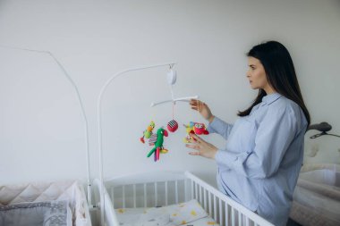 Hamile bir kadın, beyaz bir bebek beşiğinin üzerinde asılı duran bir oyuncağı dikkatlice ayarlıyor ve yeni doğan bebeğinin gelişine hazırlanıyor.