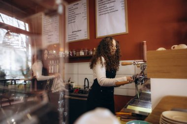 Modern bir kafede espresso makinesini çalıştıran kıvırcık saçlı genç bayan barista mükemmel müşteri hizmetleri sunuyor.