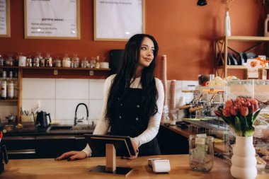 Gülümseyen kadın küçük işletme sahibi tezgahın arkasında çalışıyor, çağdaş bir kafeye bakıyor.
