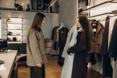 Askılardaki farklı ceketleri dikkatle inceleyen iki genç kadın şık bir giyim mağazasında yeni kıyafetler seçiyor.