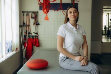 Genç bayan fizyoterapist muayene koltuğunda oturuyor ve tıbbi ekipmanlarla bir klinikte terapi seansına hazırlanıyor.