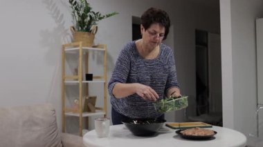 Emekli kadın evde sağlıklı bir yemek hazırlıyor, plastik bir kabın içine taze sebze ekliyor.