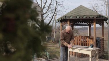 Zımpara kağıdıyla ahşap bir tahtayı zımparalayan gözlüklü yaşlı beyaz bir adam. Güneşli bir günde arka bahçesinde çalışan emekli bir zanaatkar.