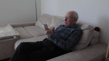 Oturma odasındaki koltukta oturan, interneti karıştıran ya da akıllı telefonunda sosyal medya kullanan yaşlı bir adam.