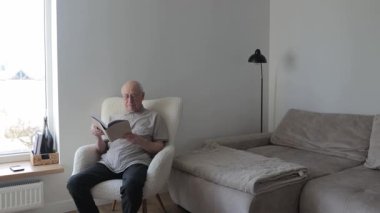 Oturma odasında kitap okumaya dalmış, parlak bir pencerenin yanındaki rahat koltukta dinlenen kıdemli beyefendi.