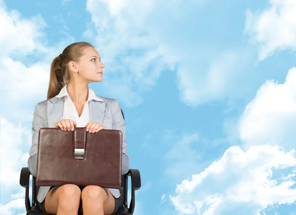 Business woman in skirt, blouse and jacket, sitting on chair. Against background of blue sky, clouds