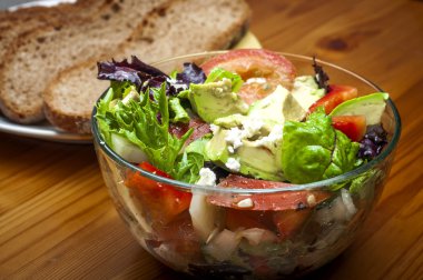 Bowl of salad with feta cheese, tomatos, avocado, leafy greens, red peppers, and onions on a wooden table with rustic sliced bread in the background
