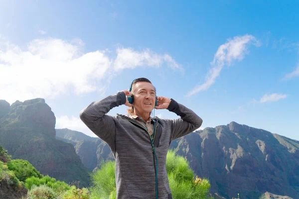 Feliz hombre maduro viajero en auriculares de pie en la cima de la colina y disfrutar de un hermoso paisaje de montaña — Foto de Stock
