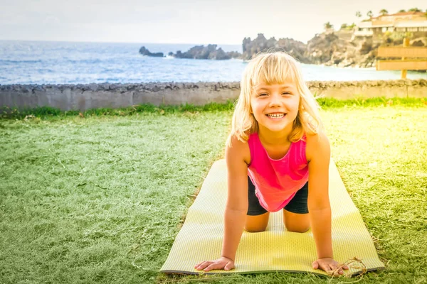 Linda niña feliz en ropa deportiva sonriendo a la cámara mientras hace ejercicio en la playa —  Fotos de Stock