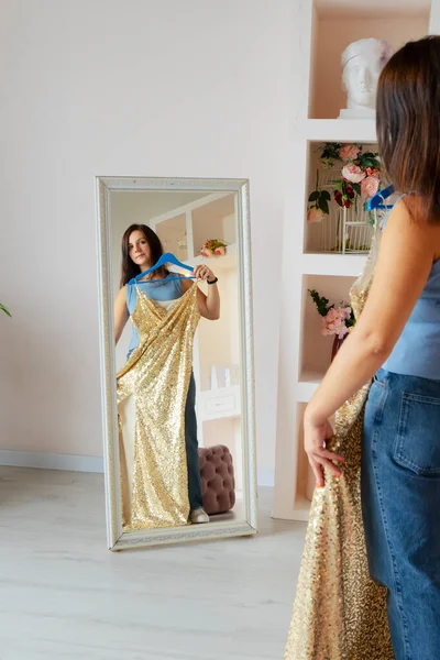 Молодая брюнетка стоит перед зеркалом дома, держа вечернее платье в золотых блёстках. — стоковое фото