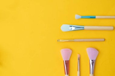 Sarı arka planda makyaj fırçaları, pastel sünger başları, tahta saplar, metalik ferruarlar, yumuşak stüdyo ışıkları, yüksek anahtarlı kompozisyon...,