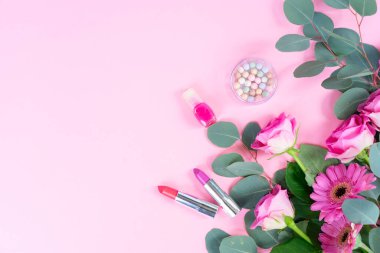 Çiçekleri ve kozmetik ürünleri olan pembe düz yatak örtüsü, yumuşak kadın havası, gül buketi ve okaliptüs dalları ruj, pudra, boncuklu mücevher ve pastelin yanında düzenlenmiş.