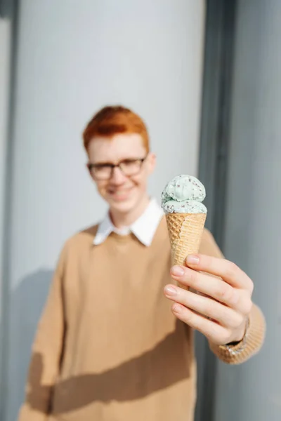 Homem de gengibre sorridente em óculos segurando cone de sorvete perto da câmera. turva — Fotografia de Stock
