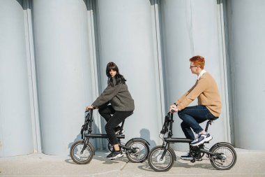 Bisiklet süren genç bir adam ve bir kadın, birbirlerine bakıyorlar. Güneşli bir sonbahar gününde. Arka planda büyük sütunlar.