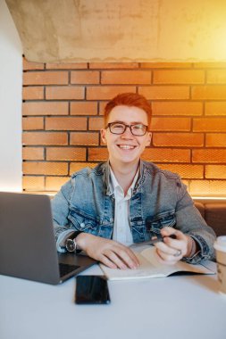 Kafedeki bir kanepede oturan, elinde kalem ve defterle kameraya bakan sevimli gülümseyen genç adam. Arkasından aydınlatılmış tuğla duvar. Kalın çerçeveli gözlük takıyor.