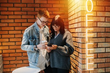 Kafede ışıl ışıl parlayan kahve tabelasıyla aydınlanmış tuğla duvarın önünde duran mutlu genç çift. İkisinin de gözlüğü kalın çerçeveli, akıllı telefon ekranına bakıyor..