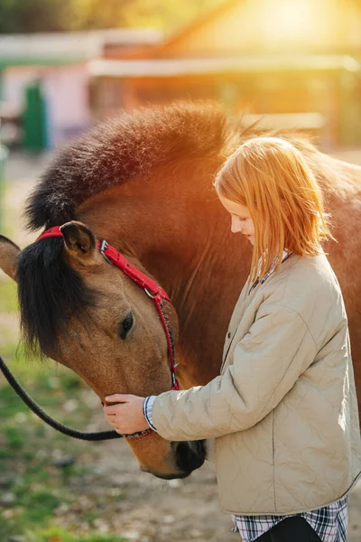 Idilic image of a preteen ginger girl petting her horse tied to wooden fence. Smiling, looking with love. Horse bowing her head. Against sunlight.