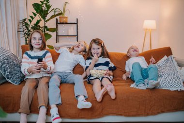 5 yaşından 10 yaşına kadar eğlenmiş, gülümseyen çocuklar bir kanepede oturur, televizyon izler, patlamış mısır yerler.
