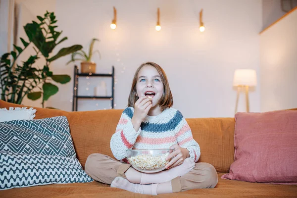 怒目而视的小女孩看电视 吃爆米花 坐在客厅的沙发上 — 图库照片
