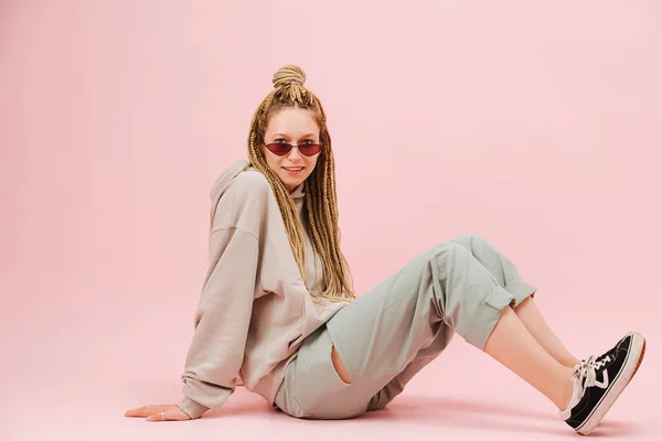 ピンクの背景の上に床に横に座ってスタイリッシュなブロンドのアフロ編組と狭いサングラスでかわいい若い女性 ゆったりとしたグレーの服とスニーカー — ストック写真