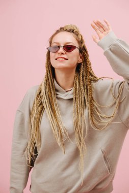 Dar güneş gözlüklü, sevimli, gülümseyen, pembe arka planda sarı afro örgülü genç bir kadın. Başını dik tut, birine bakıyor, elini sallıyor..
