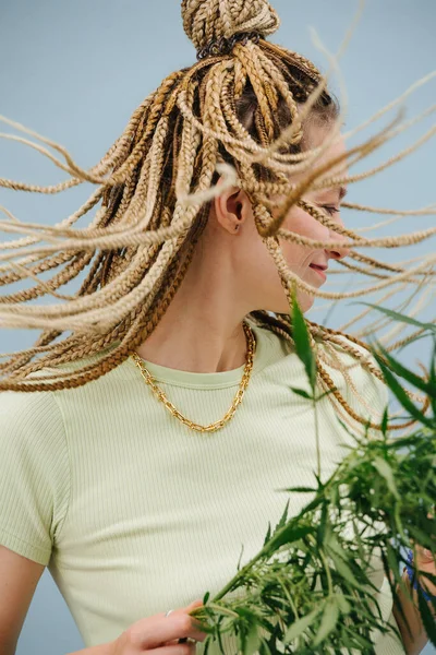 青の背景にスタイリッシュな金髪アフロ編組を持つ幸せな若い女性 彼女の髪を振って大麻の植物を手に持って — ストック写真