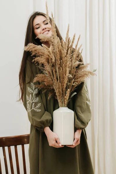 Mujer sonriente en un vestido de oliva sosteniendo jarrón con ramo de cereales en las manos — Foto de Stock
