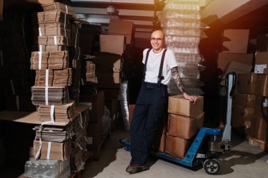 Gülümseyen erkek depo çalışanı palet kutusuna yaslanıyor.