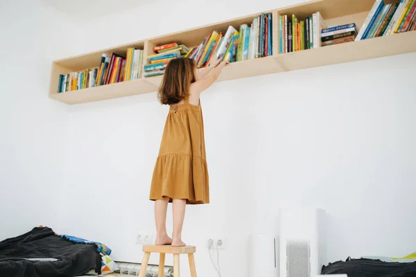 Preschool girl got up on a stool to get a book off the shelf — Fotografia de Stock